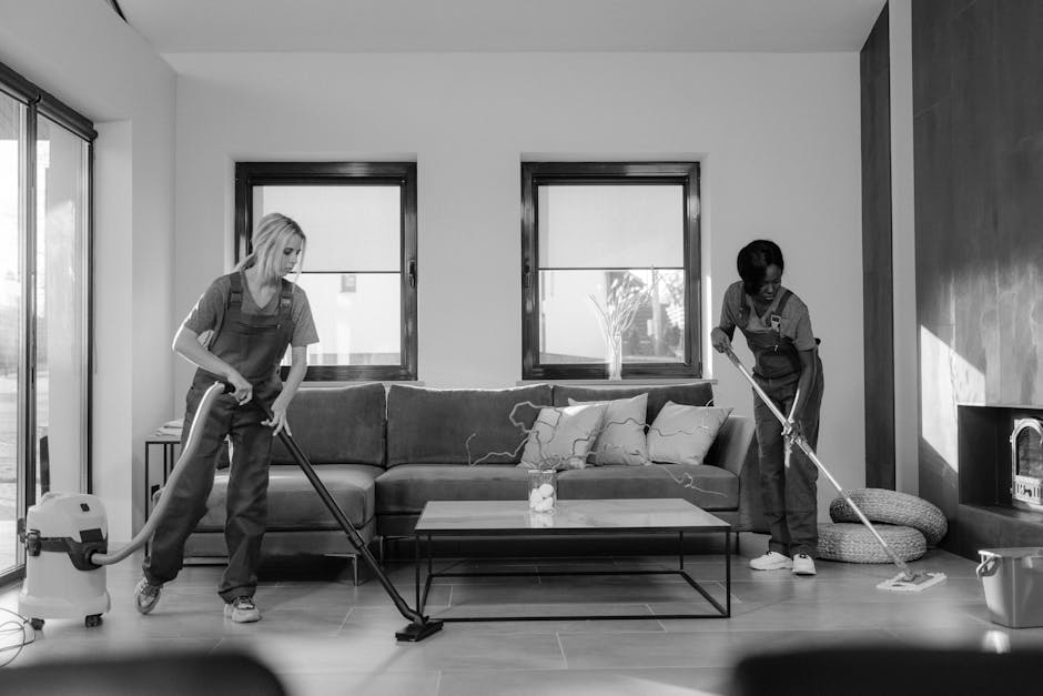 Two professional cleaners from Edgware Cleaner are performing surface cleaning and deep cleaning in a modern living room. One cleaner is using a vacuum cleaner to remove dust and debris from the hardwood floor, while the other is utilizing a mop to clean the tiled area near the fireplace. The room features a contemporary style with a grey fabric sofa, decorative cushions, and a glass coffee table centered on a wooden floor. Natural light enters through two large windows in the background, illuminating the clean and tidy space. The scene demonstrates thorough domestic cleaning and sanitation efforts to maintain a hygienic living environment.