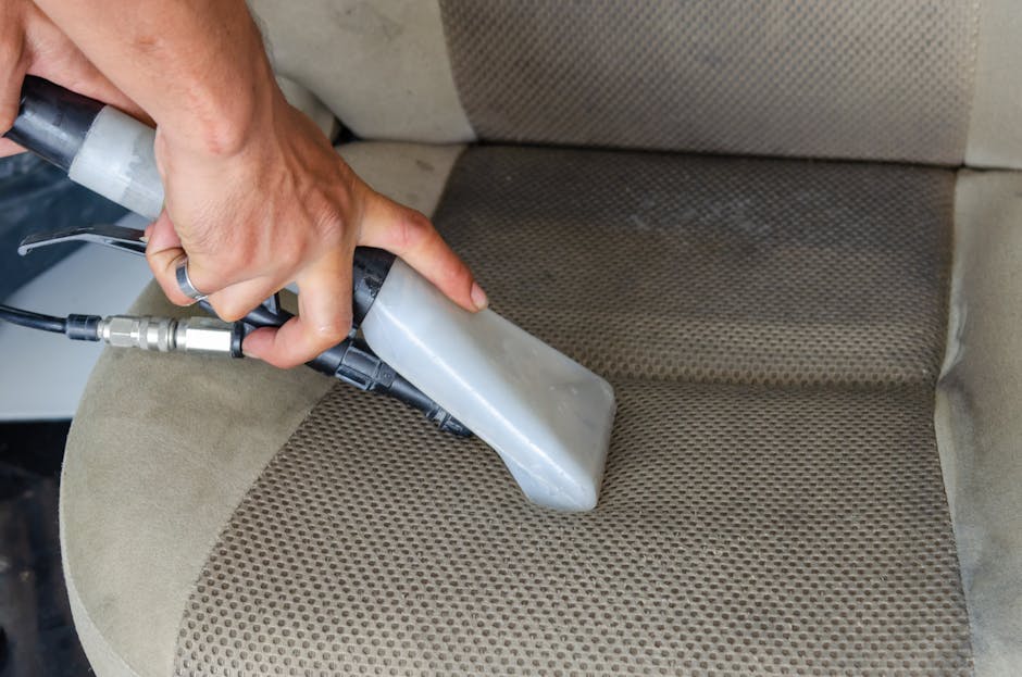 A close-up of a person using a handheld upholstery cleaning machine on a beige fabric upholstered chair in a room with natural light. The device has a gray and black body with a hose connected to a cleaning solution supply, and the nozzle is pressed against the textured fabric, removing dirt and stains. The chair's armrest is visible in the background, and the surrounding area appears clean and well-maintained. The scene exemplifies professional upholstery surface cleaning and maintenance, showcasing detailed fabric cleaning processes as part of a domestic cleaning service from Edgware Cleaner, highlighting their upholstery deep cleaning expertise.