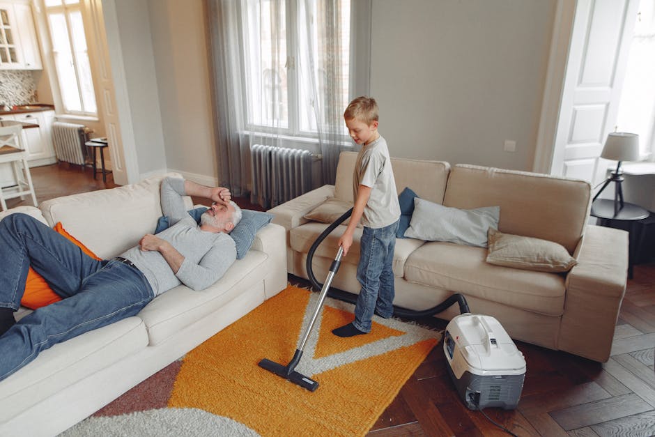 A young boy operating a vacuum cleaner on a colorful orange and beige patterned rug in a living room, cleaning the wooden floor near a beige sofa. An older man with gray hair and a beard, dressed in a gray sweater and blue jeans, is resting on the sofa, appearing relaxed and comfortable. The room features large windows with sheer curtains allowing natural light in, a radiator beneath the window, and several throw pillows on the sofa. The setting highlights domestic cleaning and surface maintenance, with the vacuum cleaner and the clean, polished floors emphasizing hygiene and deep cleaning, all under soft, natural lighting. Edgware Cleaner is mentioned as the provider of professional domestic cleaning services.