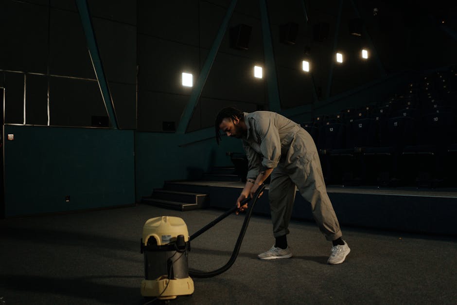 A person in a grey jumpsuit and sneakers is using a vacuum cleaner to deep clean a carpeted floor in a vacant auditorium with dark walls, steps leading up to a stage, and overhead lighting. The room is dimly lit, emphasizing the cleanliness of the carpet surface. The individual appears focused on the surface cleaning process, highlighting hygiene and maintenance practices. The scene showcases professional cleaning equipment in use within a large indoor space, reflecting thorough carpet sanitisation effort expected from Edgware Cleaner, a specialist in domestic and commercial cleaning services, as referenced on edgwarecleaner.co.uk.