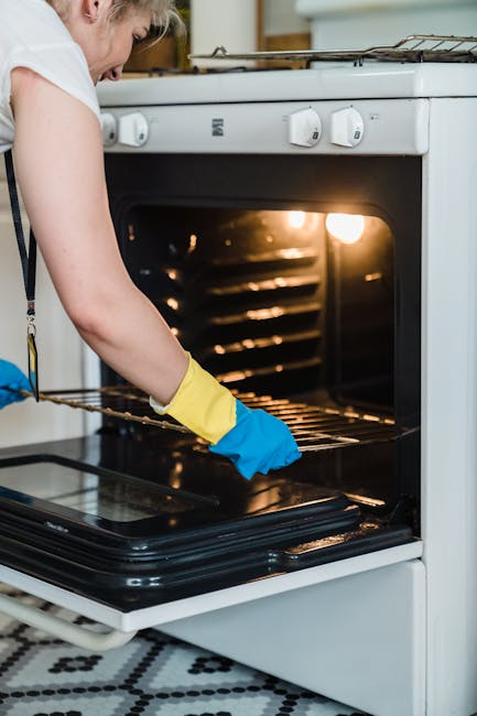 A person wearing blue and yellow rubber gloves is holding a green spray bottle and a cleaning cloth, performing surface cleaning on a white oven stovetop with black metal grates in a kitchen. The stov